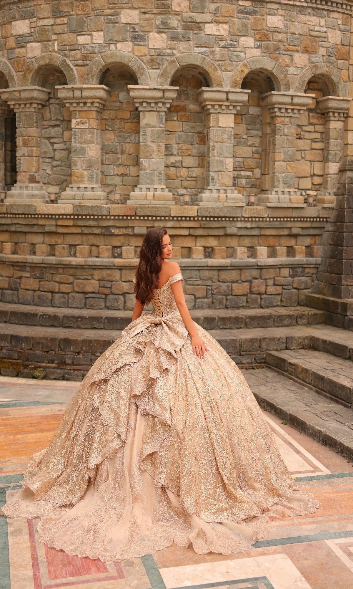 A woman in the Quinceanera Dress 54324 By Amarra, an ornate off-the-shoulder gold gown with lace embroidery, stands on a patterned stone floor before a historic building with arched windows.